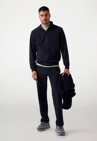 Veste de survêtement zippée bleu marine et pantalon assorti en tissu doux, assortis à un sous-vêtement blanc. Le mannequin tient un pull foncé, portant des baskets grises.