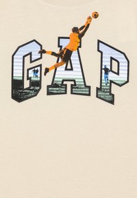 Beige T-krekls ar izteiksmīgu "GAP" dizainu. Grafika ietver lecošu vārtsargu un divus spēlētājus, ar izteiksmīgām krāsām un tekstūrām.