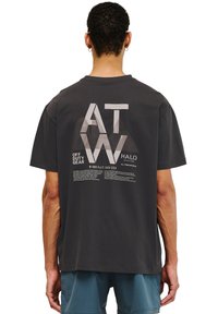 Mörkgrå t-shirt av bomull med en grafisk design med bokstäverna "ATW" i ljusgrå och textdetaljer på baksidan. Korta ärmar.