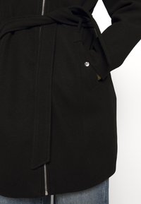 Zwarte wollen jas met een riem in de taille, een ritssluiting aan de voorkant, zijzakken en een subtiele textuur. Verfraaid met een glanzende knoopdetail op de zak.