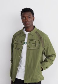 Ελαφρύ ελαιόχρωμο μπουφάν με μαύρη περιγραμματική λογότυπο, κλείσιμο με κουμπί και δικτυωτές πλευρικές πάνελ, στερεωμένο πάνω από ένα λευκό μπλουζάκι.