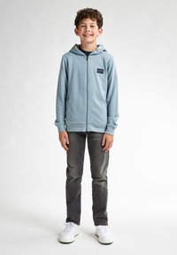 Ljusblå zip-up huva med dragsko, sidofickor och en liten logotypapplikation, parat med grå jeans och vita sneakers.