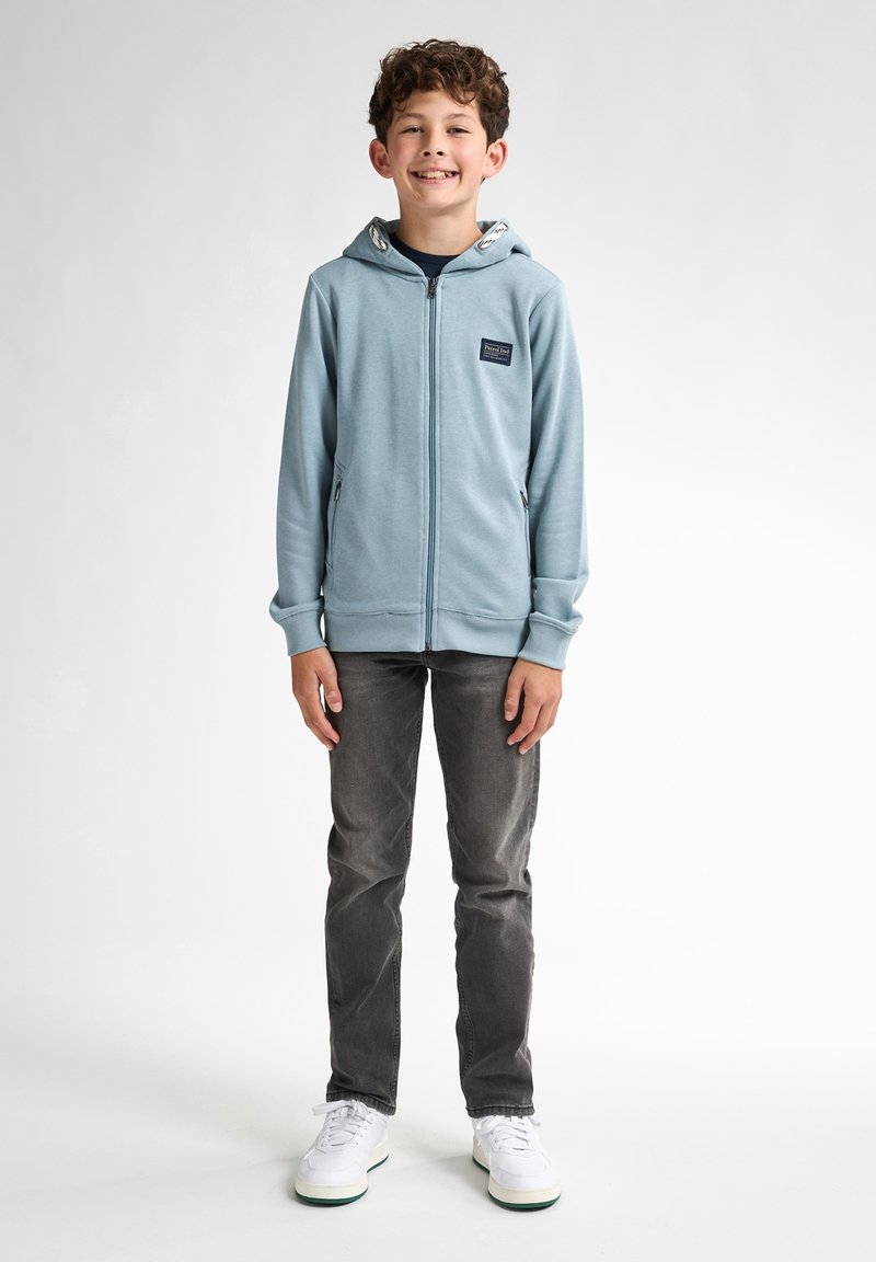 Ljusblå zip-up huva med dragsko, sidofickor och en liten logotypapplikation, parat med grå jeans och vita sneakers.