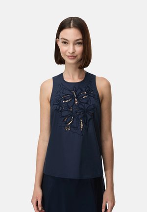 Donna con taglio capelli bob castano liscio che indossa una canotta blu scuro con ricamo floreale traforato sul petto, in piedi davanti a uno sfondo semplice.