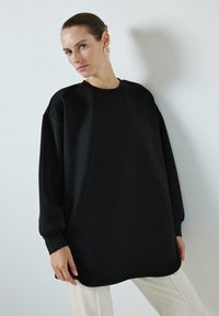 Crni oversized dukserica s okruglim izrezom i uskim dugim rukavima. Glatka tkanina, suptilan sjaj i zaobljeni rub.