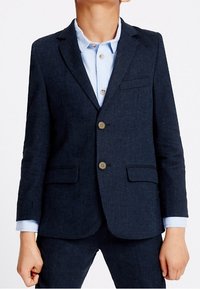 Granatowa marynarka wykonana z teksturowanej tkaniny, z dwiema guzikami, przednimi kieszeniami i wykładanym kołnierzykiem, w połączeniu z jasnoniebieską koszulą.