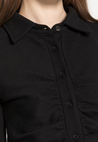 Svart skjorta med knappar och ribbat mönster, med krage och sju svarta knappar, visar svag rynkning framme.