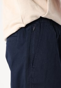 Pantaloni blu navy con vita elasticizzata, tasche laterali e una leggera lucentezza. Il tessuto appare liscio con una vestibilità comoda, evidenziando i dettagli delle cuciture.