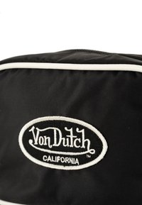 Sac en nylon noir avec un biais blanc, présentant un logo ovale brodé avec "Von Dutch" et "CALIFORNIA" en lettres cursives et en capitales.