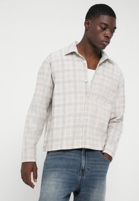 Mlad muškarac u svijetlosmeđem kariranom košulji s dugim rukavima preko bijele majice, u plavim trapericama, pozira s rukama u džepovima ili blizu njih, gledajući dolje.