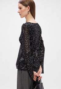 Μαύρη διάφανη μπλούζα με ασημένια σχέδια σε σπείρα, μακριά μανίκια και χαλαρή εφαρμογή. Συνδυασμένη με γκρι παντελόνι με ρίγες και μαύρη τσάντα.