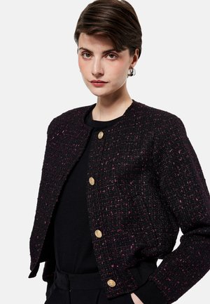 Jonge vrouw met kort bruin haar, die een zwarte gestructureerde jas met gouden knopen draagt over een zwart topje, staand tegen een effen achtergrond.