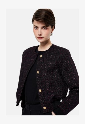 Jonge vrouw met kort bruin haar, die een zwarte gestructureerde jas met gouden knopen draagt over een zwart topje, staand tegen een effen achtergrond.