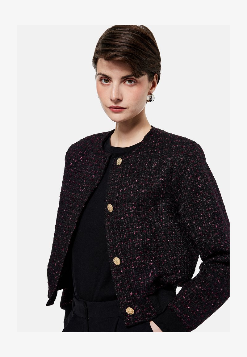 Jeune femme aux cheveux courts bruns portant une veste noire texturée avec des boutons dorés par-dessus un haut noir, debout devant un fond uni.