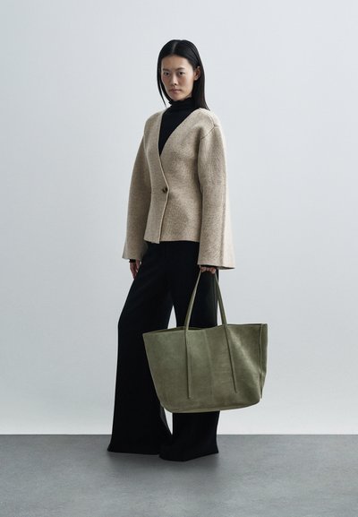 Beige gebreid vest van een wolblend met een knoop, zwarte coltrui, wijde zwarte broek en een grote groene suède totebag met lange hengsels.