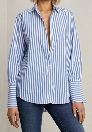 Vrouw draagt een blauw-wit verticale gestreept overhemd met lange mouwen en donkere blauwe spijkerbroek, staand tegen een effen achtergrond.