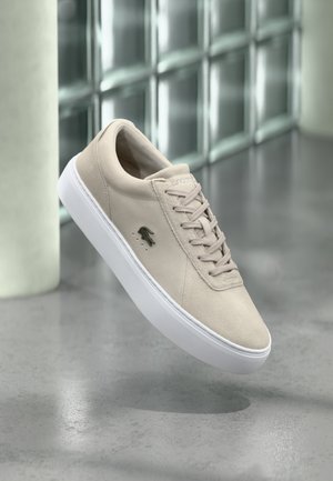 Baskets en daim beige avec semelle blanche, lacets et petit logo crocodile sur le côté, flottant au-dessus d'un sol en béton poli.