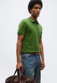 Mann mit grünem Poloshirt und blauen Jeans, der eine braune Ledertasche vor einem hellen, schlichten Hintergrund hält.