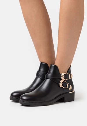 Bottines en cuir noir avec un faible talon, ornées de deux boucles dorées et d'une texture lisse.