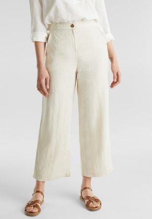 Frau trägt hochtaillierte, weit geschnittene cremefarbene Hosen mit Knopfverschluss, kombiniert mit braunen, flachen Riemchensandalen und einem weißen Hemd mit hochgekrempelten Ärmeln.