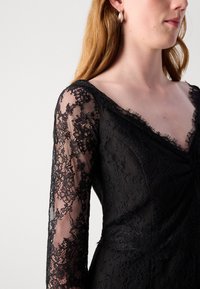 Robe en dentelle noire avec des manches longues et transparentes, ainsi qu'un décolleté en V. Texturée avec des motifs floraux complexes et des détails en franges délicates.