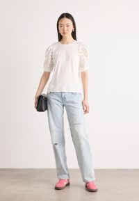 Un tricou alb cu manșete din dantelă, asociat cu o pereche de blugi ușor destrămați de culoare bleu deschis și adidași roz, ținând o geantă mică de umăr neagră.