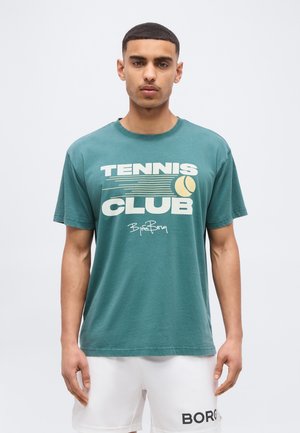 Zöld "TENNIS CLUB" feliratú pólót és fehér rövidnadrágot viselő férfi, egyszínű világos háttér előtt állva.