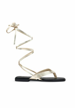 Sandalias planas con suela negra y tiras metálicas doradas. Las tiras se envuelven alrededor del tobillo en un diseño en espiral, con una textura suave y brillante.