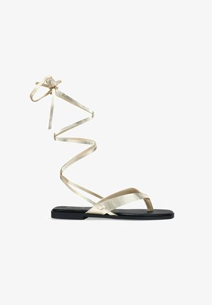 Sandalias planas con suela negra y tiras metálicas doradas. Las tiras se envuelven alrededor del tobillo en un diseño en espiral, con una textura suave y brillante.