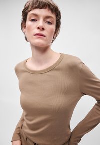 Καφετί μπλούζα με μακριά μανίκια, φτιαγμένη από ραβδωτό υλικό, με φαρδύ στρογγυλό ντεκολτέ και εφαρμοστή σιλουέτα, φορεμένη με τα χέρια στη μέση.