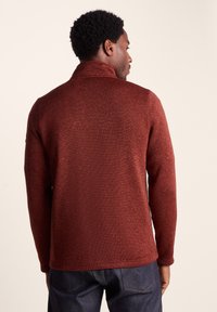 Rust-colored sweater with a textured knit, high collar, long sleeves, and a straight fit, paired with dark denim jeans.