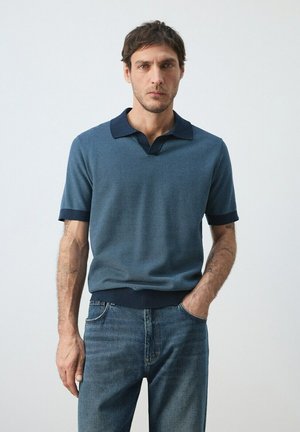 Hombre de cabello oscuro vistiendo un polo de punto azul de manga corta y jeans, de pie con una mano en el bolsillo contra un fondo claro y liso.