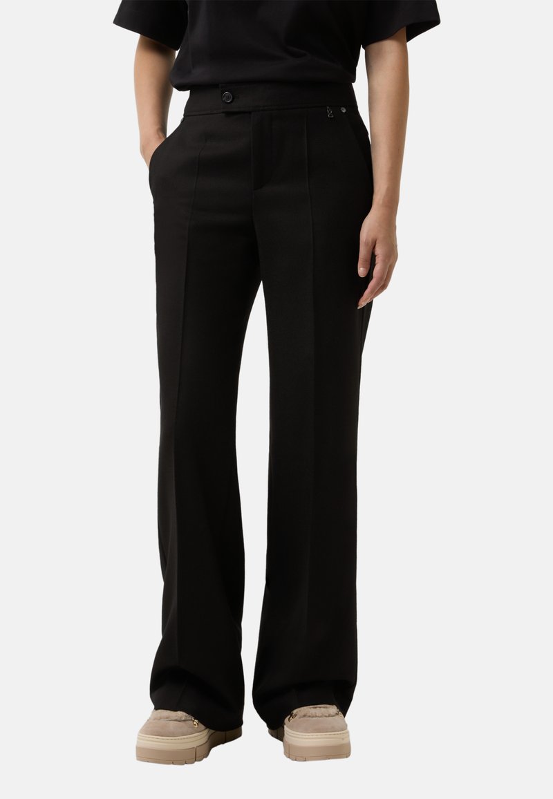 Žena oblečená v černých kalhotách s vysokým pasem a širokými nohavicemi s předními záhyby a béžovými platformovými slip-on botami na hladkém pozadí.