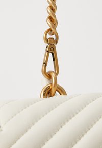 Baltas quiltots ādas rokassoma ar zelta ķēdes siksnu un sprādzi. Iezīmēta ar apaļu metāla gredzenu un iecirstu zīmola nosaukumu uz piederumiem.