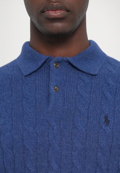 Blå strikket polo med kabelmønster, med klassisk krage og to brune knapper. Logo brodert i svart på brystet.