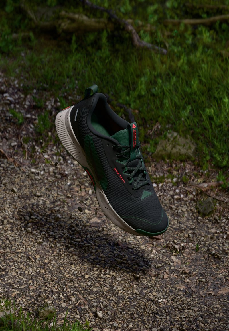 Chaussure de trail running noire et verte avec une tige en maille texturée, un collier rembourré et une semelle extérieure résistante à la boue. Présente des accents rouges et des détails réfléchissants.