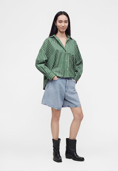 Jeune femme portant une chemise rayée verte, un short en jean clair jusqu'aux genoux et des bottes noires mi-mollet, mains dans les poches.