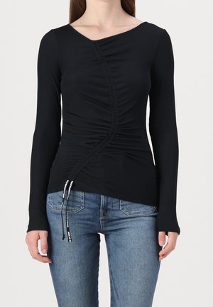 Vrouw draagt een aansluitende zwarte top met lange mouwen met geplooide details en verstelbare trekkoorden, gecombineerd met een blauwe spijkerbroek.