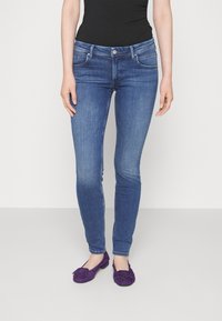 Vaqueros ajustados de mezclilla de lavado medio, con bolsillos frontales y un sutil desvanecimiento, acompañados de zapatos planos de ante morados.
