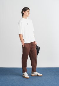 Vit oversized t-shirt med logotyp, bruna sweatpants med logotyp på benet och vita sneakers med svarta detaljer, stående på blå matta.