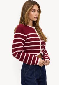 Cárdigan corto de rayas rojas y rosas, textura de punto, con botones redondos dorados en la parte delantera y mangas largas. Llevado sobre pantalones azul oscuro.