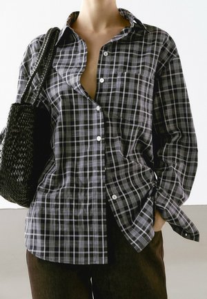 Personne portant une chemise à carreaux noire et blanche boutonnée, une main dans la poche, portant un sac d'épaule noir tissé.