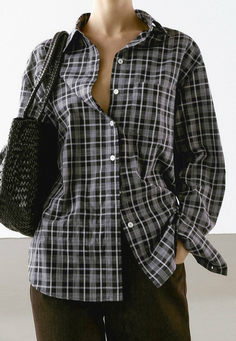 Personne portant une chemise à carreaux noire et blanche boutonnée, une main dans la poche, portant un sac d'épaule noir tissé.