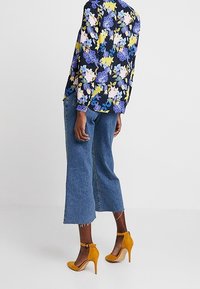 Blůza s květinovým vzorem v námořnické modři s žlutými, modrými a růžovými květy; v kombinaci s světle modrými zkrácenými džíny a hořčicově žlutými vysokými podpatky.