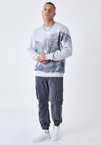 Lichtegrijze sweatshirt met een vleugeld ontwerp; gecombineerd met donkergrijze cargobroek met zijzakken; witte sneakers.