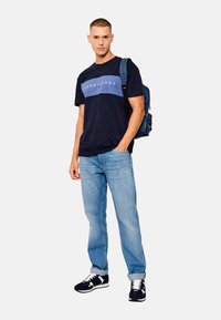 Camiseta de marina con una franja horizontal azul claro y el logo "TOMMY JEANS", combinada con jeans azul claro y zapatillas oscuras. Mochila al hombro.