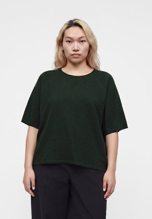Jeune personne aux longs cheveux blonds portant une chemise à manches courtes vert foncé et un pantalon noir sur un fond clair uni.