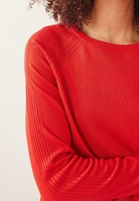 Pull rouge avec détails côtelés sur les manches et l'encolure ; texture douce et coupe ajustée, mettant en valeur une finition de tissu lisse.