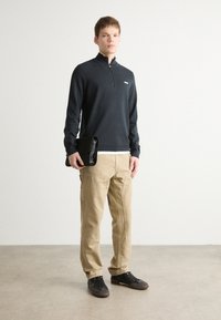 Suéter oscuro de manga larga con cuello con cremallera, pantalones cargo beige y zapatillas negras. El modelo sostiene un bolso de mano negro. Fondo de suelo de concreto.