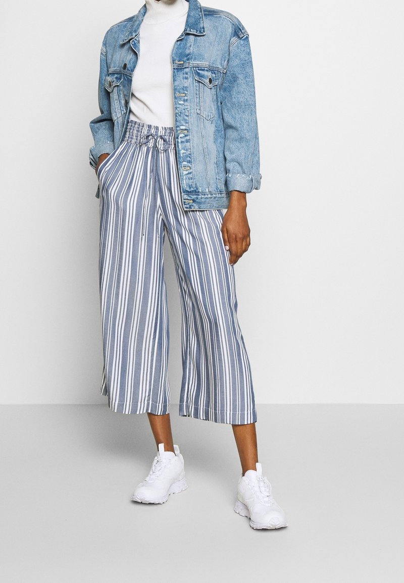 Personne portant une veste en jean, un col roulé blanc, un pantalon large rayé bleu et blanc coupé au-dessus de la cheville, et des baskets blanches, sur un fond uni.